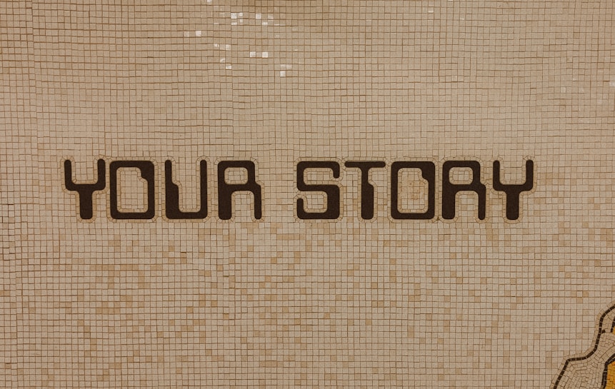 The image contains a mosaic wall with the words 'YOUR STORY' written in bold, dark letters. The tiles are small and mostly uniform, giving a textured appearance to the background.