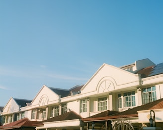 A row of suburban houses featuring classic architecture with triangular gables, large windows, and brown tiled roofs. The houses are well-maintained and the sky above is clear and blue.