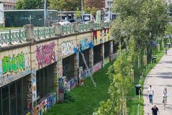 A long wall covered in vibrant graffiti art runs alongside a walkway. The wall features various styles and colors of street art, suggesting a creative urban environment. A few people walk and cycle along the path lined with newly planted trees, indicating an effort to green the area. The atmosphere combines art and nature in an urban setting.