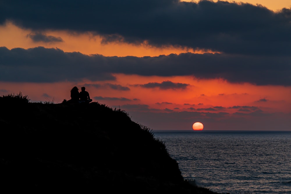 silhouette of 2 people sitting on rock formation during sunset