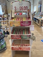 A wooden display stand in a store showcasing various Cottage Delight products. The top sign advertises a promotion of 3 items for £6.00. There are multiple shelves filled with jars of condiments, each shelf labeled with descriptive tags indicating different flavors such as all butter lemon curd, zingy raspberry jam, tomato, garlic & ginger chutney, and English country cider chutney. The store appears well-organized, and there are other products visible in the background.