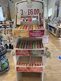 A wooden display stand in a store showcasing various Cottage Delight products. The top sign advertises a promotion of 3 items for £6.00. There are multiple shelves filled with jars of condiments, each shelf labeled with descriptive tags indicating different flavors such as all butter lemon curd, zingy raspberry jam, tomato, garlic & ginger chutney, and English country cider chutney. The store appears well-organized, and there are other products visible in the background.
