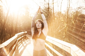 A person with tattoos stands on a wooden bridge, arms raised above the head in a yoga pose, with the sun shining brightly through the trees, creating a warm, golden glow and long shadows across the path.