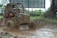 A small off-road vehicle is driving through a muddy terrain, splattering mud around as it moves. The driver is wearing protective gear and a helmet, indicating a rugged outdoor activity. The background shows a fenced area with greenery and a billboard.