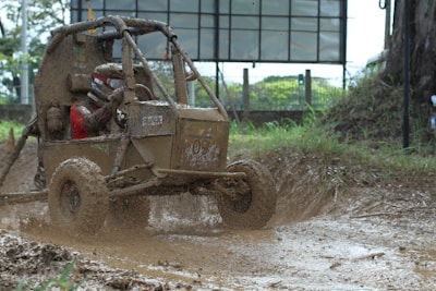 A small off-road vehicle is driving through a muddy terrain, splattering mud around as it moves. The driver is wearing protective gear and a helmet, indicating a rugged outdoor activity. The background shows a fenced area with greenery and a billboard.