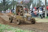 A muddy off-road buggy with two occupants, both wearing helmets and protective gear. They are navigating a dirt track during a racing event. The crowd in the background is observing from behind barriers, and there are trees and some flags visible.