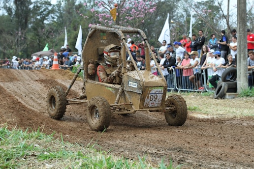 A muddy off-road buggy with two occupants, both wearing helmets and protective gear. They are navigating a dirt track during a racing event. The crowd in the background is observing from behind barriers, and there are trees and some flags visible.