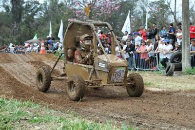 A muddy off-road buggy with two occupants, both wearing helmets and protective gear. They are navigating a dirt track during a racing event. The crowd in the background is observing from behind barriers, and there are trees and some flags visible.