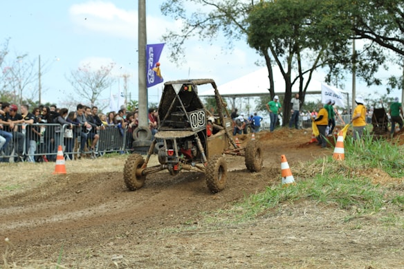A muddy off-road vehicle is speeding through a dirt track, kicking up mud as it goes, with orange and white traffic cones marking the course. In the background, a crowd of spectators stands behind metal barriers, observing the event, while a few trees and a tent are visible.