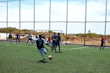 A soccer match is taking place on a field with players actively engaged in the game. One player wearing a black and white jersey with the number 7 is about to kick the ball. Opponents in blue and orange bibs are positioned near the goal, and a referee dressed in red observes the action from the side.