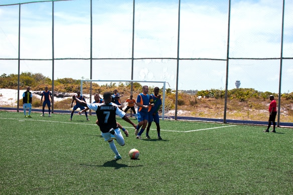 A soccer match is taking place on a field with players actively engaged in the game. One player wearing a black and white jersey with the number 7 is about to kick the ball. Opponents in blue and orange bibs are positioned near the goal, and a referee dressed in red observes the action from the side.