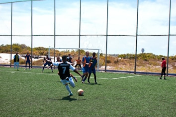 A soccer match is taking place on a field with players actively engaged in the game. One player wearing a black and white jersey with the number 7 is about to kick the ball. Opponents in blue and orange bibs are positioned near the goal, and a referee dressed in red observes the action from the side.