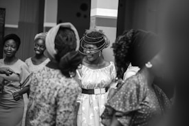 A group of women stand together, engaged in conversation and smiling, some are wearing intricately designed outfits with headscarves. The setting appears to be indoors with soft lighting and a relaxed atmosphere.