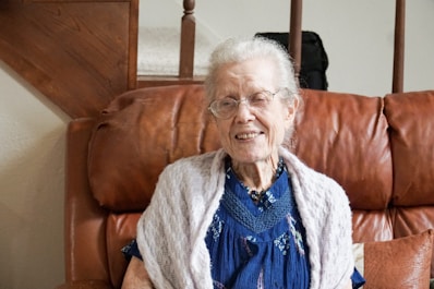 An elderly woman with white hair and glasses is sitting comfortably on a brown leather couch. She is wearing a blue dress with a lace shawl around her shoulders. The setting includes a wooden staircase in the background, suggesting a cozy home environment.