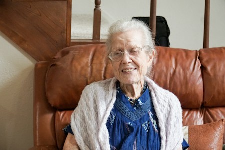 An elderly woman with white hair and glasses is sitting comfortably on a brown leather couch. She is wearing a blue dress with a lace shawl around her shoulders. The setting includes a wooden staircase in the background, suggesting a cozy home environment.