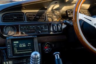 The interior of a vintage car features a retro dashboard with wooden elements. Analog gauges, a classic air vent design, and a wooden steering wheel are prominently displayed. The dashboard includes a digital display showing AFR with current, minimum, and maximum values. The gear shift lever and other control knobs are also visible.