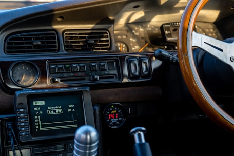 The interior of a vintage car features a retro dashboard with wooden elements. Analog gauges, a classic air vent design, and a wooden steering wheel are prominently displayed. The dashboard includes a digital display showing AFR with current, minimum, and maximum values. The gear shift lever and other control knobs are also visible.