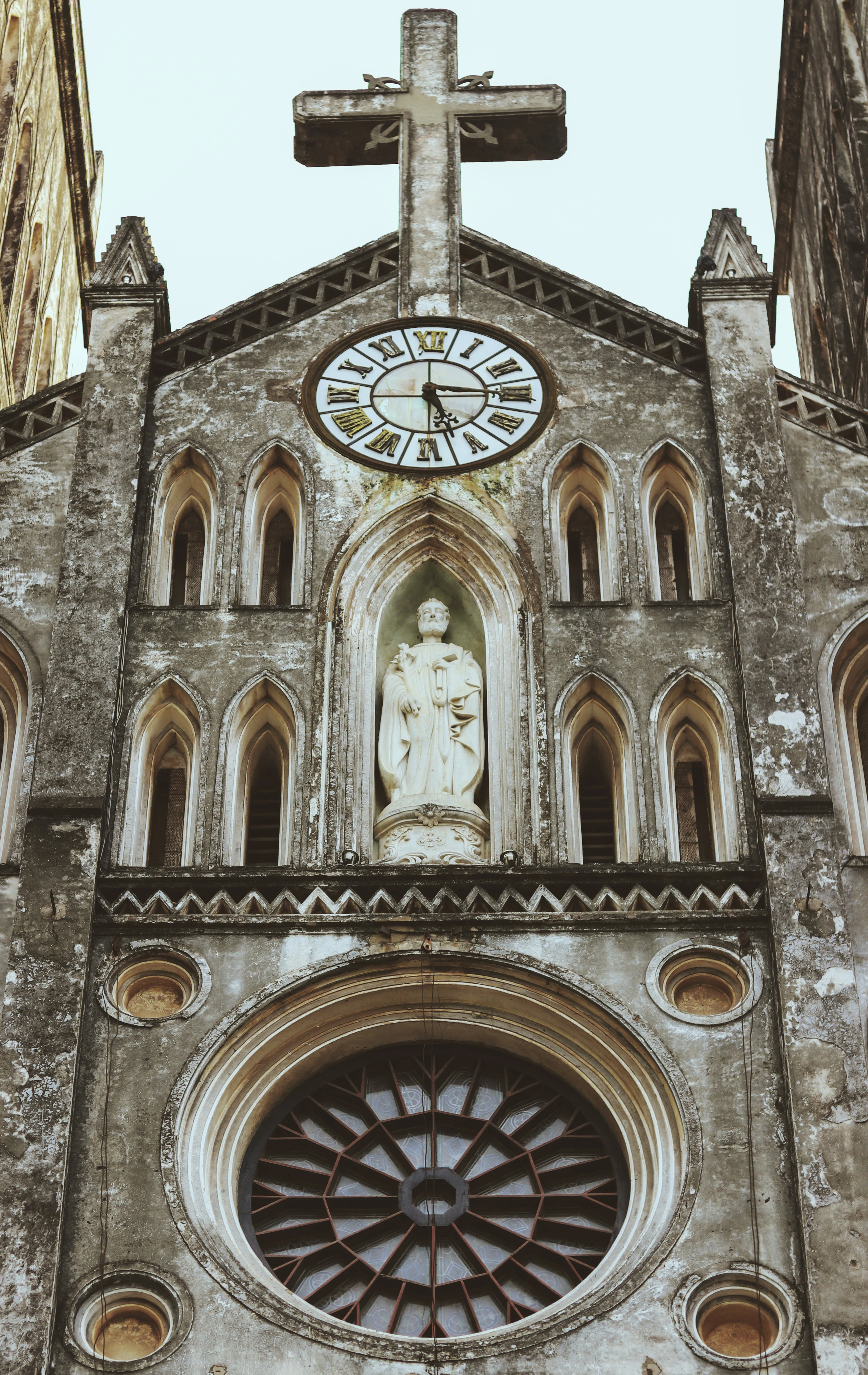 The weathered grey neo-Gothic facade of St Joseph's Cathedral in Hanoi seen from below, showing its stone cross, clock face, a saint's statue in an arched niche and the spoked rose window beneath.
