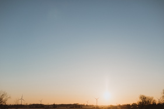 A serene landscape at sunrise or sunset with the sun near the horizon. Wind turbines are scattered across a vast plain, with trees framing the scene at the edges. The sky transitions smoothly from a pale blue at the top to warm yellow and orange hues near the horizon.