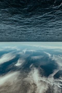 An inverted image where the ocean appears at the top and a cloudy sky follows beneath. The water exhibits a dark, textured surface, while the sky features various layers of white and gray clouds, creating an illusion of the world turned upside down.