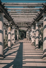 A long, sunlit pergola with a series of evenly spaced stone columns supporting a wooden lattice. Shadows from the lattice create intricate patterns on the paved walkway below. Greenery lines the sides of the path, and at the far end, more lush vegetation is visible.