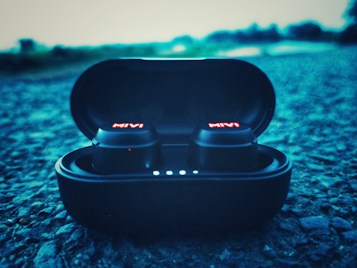 A pair of wireless earbuds in a charging case are placed on a textured surface, with the brand name glowing in red on the earbuds. The background appears blurred, giving a sense of depth and focus on the earbuds.
