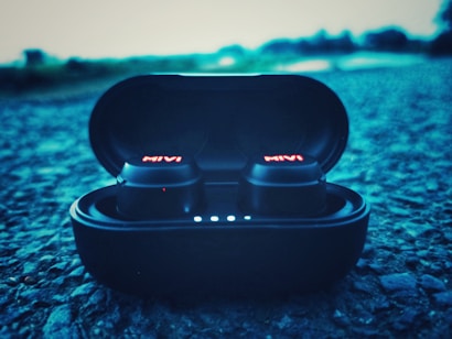 A pair of wireless earbuds in a charging case are placed on a textured surface, with the brand name glowing in red on the earbuds. The background appears blurred, giving a sense of depth and focus on the earbuds.