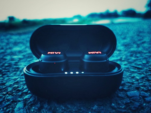 A pair of wireless earbuds in a charging case are placed on a textured surface, with the brand name glowing in red on the earbuds. The background appears blurred, giving a sense of depth and focus on the earbuds.