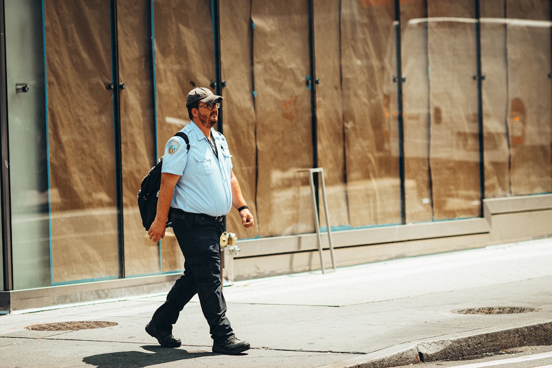 man in blue polo shirt and black pants wearing black hat standing on sidewalk during daytime