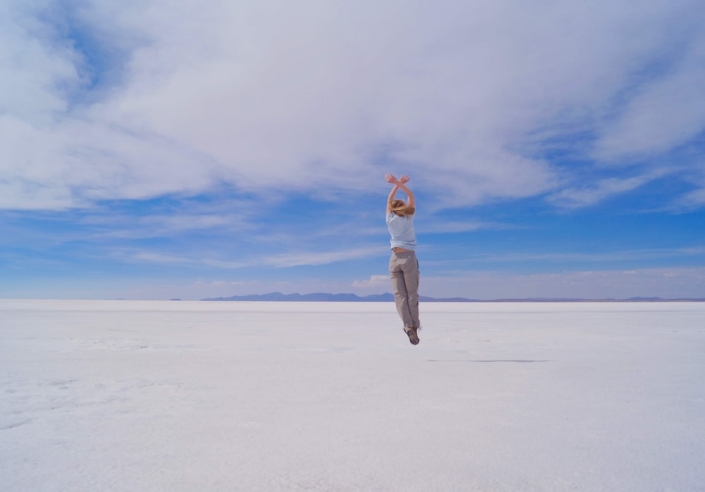 mulher na jaqueta branca e calças cinzentas de pé na areia branca sob o céu azul durante o dia