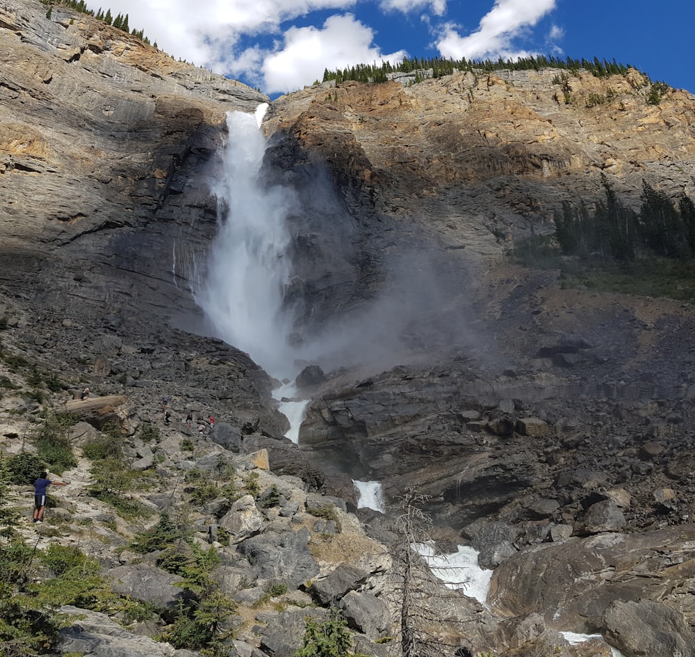 person in blue jacket standing on rock formation near waterfalls during daytime