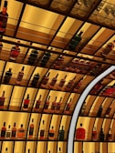 A series of shelves stocked with various bottles of alcoholic beverages, primarily featuring different types of liquor such as whiskey, vodka, and rum. The shelves are illuminated with a warm yellow light, creating an inviting ambiance. The arrangement of the bottles is neat and organized, with some sections having a repeating pattern of the same bottle.