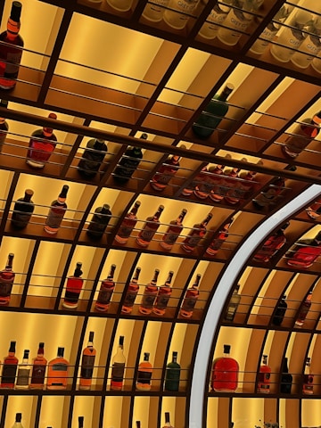 A series of shelves stocked with various bottles of alcoholic beverages, primarily featuring different types of liquor such as whiskey, vodka, and rum. The shelves are illuminated with a warm yellow light, creating an inviting ambiance. The arrangement of the bottles is neat and organized, with some sections having a repeating pattern of the same bottle.