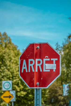 A red octagonal stop sign with the word 'ARR&Ecirc;T' is displayed prominently against a clear blue sky. The sign shows signs of wear, evident by a small patch of covering material. Trees with green foliage are visible in the background, along with other road signs that are slightly blurred.