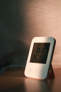 A digital thermometer or clock with a rounded rectangular shape stands on a flat wooden surface. The device displays numbers and has buttons labeled 'SET', '+', and '-'. The lighting creates a warm ambiance with shadows cast on the background.