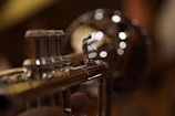 A close-up of a shiny brass trumpet with blurred circular lights in the background creating a bokeh effect. The foreground shows detailed parts of the trumpet, such as the valves and tubing, with reflections highlighting the polished metal surface.