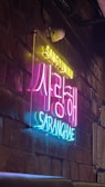 Neon signs display Korean and English text against a textured brick wall, creating a vibrant and artistic ambience. The sign has bright, contrasting colors with an industrial interior design setting.