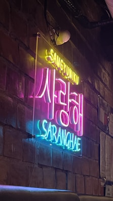 Neon signs display Korean and English text against a textured brick wall, creating a vibrant and artistic ambience. The sign has bright, contrasting colors with an industrial interior design setting.