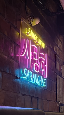 Neon signs display Korean and English text against a textured brick wall, creating a vibrant and artistic ambience. The sign has bright, contrasting colors with an industrial interior design setting.