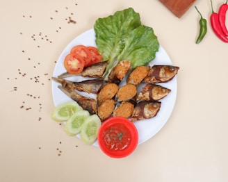 A neatly arranged dish with a variety of foods on a white plate. There are fried fish stuffed with a spiced filling, slices of fresh tomato and cucumber, and leafy greens. A small red bowl contains a sauce, possibly tomato-based, and the plate is adorned with scattered coriander seeds. Red and green chili peppers, along with a wooden block, are also visible around the plate.