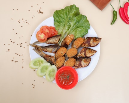 A neatly arranged dish with a variety of foods on a white plate. There are fried fish stuffed with a spiced filling, slices of fresh tomato and cucumber, and leafy greens. A small red bowl contains a sauce, possibly tomato-based, and the plate is adorned with scattered coriander seeds. Red and green chili peppers, along with a wooden block, are also visible around the plate.