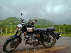 A classic motorcycle is parked on a wet road with lush green hills in the background under a cloudy sky. The motorbike features chrome details and a logo on the tank.