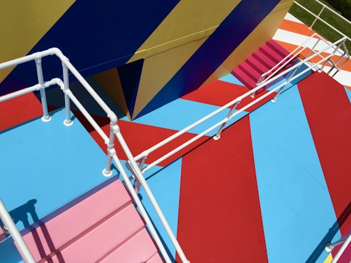 A vibrantly painted outdoor staircase features bold geometric patterns and colors. The white railing contrasts with the vivid red, blue, yellow, and pink floor sections. The angular design creates a visually striking perspective.