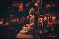 A dimly lit scene featuring a metallic statue of Buddha sitting in a serene pose. The background includes a Tuk-Tuk vehicle adorned with lights and a sign with text in Thai script. The overall ambiance is warm with an emphasis on reflective surfaces and intricate patterns.