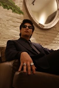 A person wearing sunglasses and a dark suit is seated on a leather couch in an upscale, dimly-lit room. Their hand, adorned with two prominent rings, is resting confidently on the armrest. A brick wall is in the background, featuring a large, ornate mirror that partially reflects the setting.