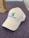 A white baseball cap with the words 'Voices of Autism' embroidered in green alongside a logo featuring a stylized person and airplane. The cap is placed on a textured, purple surface, with part of a brown paper bag visible in the background.