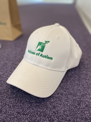 A white baseball cap with the words 'Voices of Autism' embroidered in green alongside a logo featuring a stylized person and airplane. The cap is placed on a textured, purple surface, with part of a brown paper bag visible in the background.
