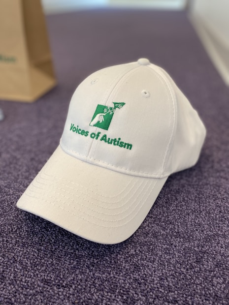 A white baseball cap with the words 'Voices of Autism' embroidered in green alongside a logo featuring a stylized person and airplane. The cap is placed on a textured, purple surface, with part of a brown paper bag visible in the background.