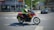 A person, wearing a helmet and a high-visibility vest, rides a red motorcycle across an urban street. The motorbike is equipped with side panniers, and there is a car parked on the side of the road. Buildings and a small number of trees are visible in the background under clear skies.