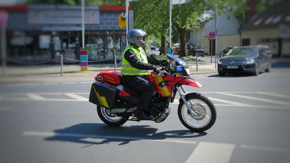 A person, wearing a helmet and a high-visibility vest, rides a red motorcycle across an urban street. The motorbike is equipped with side panniers, and there is a car parked on the side of the road. Buildings and a small number of trees are visible in the background under clear skies.